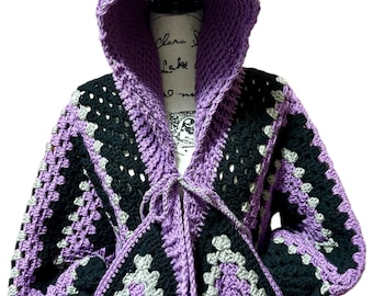 May include: A handmade crochet hooded jacket in shades of purple, black, and gray. The jacket features a hood, long sleeves, and a tie closure. The design incorporates granny squares for a unique, textured look. The jacket is displayed on a mannequin.