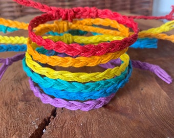 May include: A set of six colorful braided bracelets in rainbow colors, including red, orange, yellow, green, blue, and purple. The bracelets are made of a woven material and are stacked on top of each other.
