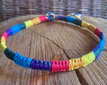 May include: A colorful woven bracelet with yellow, red, blue, purple, and black stripes. The bracelet is made of a thick, braided cord and is tied in a circle.
