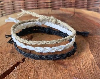 May include: Three braided bracelets in black, white, and natural colors. The bracelets are made of a woven rope material and are stacked on top of each other.