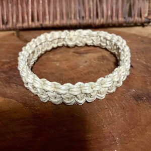 May include: A white woven bangle bracelet made of natural fibers. The bracelet is round and has a simple design.