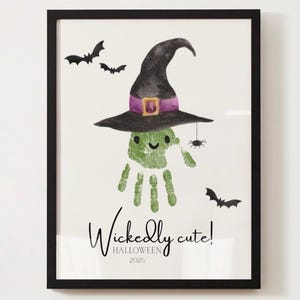 May include: A framed Halloween print. It features a green handprint with a witch's hat, a spider, and bats. The text "Wickedly cute! Halloween 2025" is written below the handprint. The artwork is in a black frame.