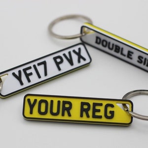 May include: Three yellow and white car registration plate keyrings. One keyring says "YOUR REG" in black letters. Another keyring says "YFI7 PVX" in black letters. The third keyring says "DOUBLE SIDE" in black letters.