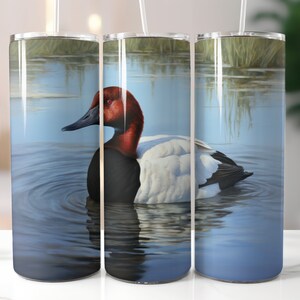 May include: A stainless steel tumbler with a design of a red-headed duck swimming in a blue lake. The duck has a white breast and black back. The lake is surrounded by green reeds.