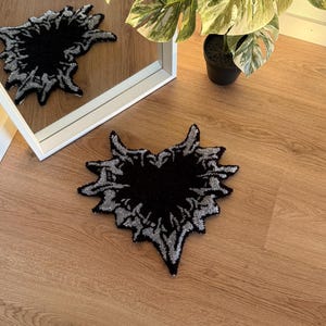 Op de afbeelding: Een zwart en zilverkleurig tapijt met een abstract ontwerp, dat lijkt op een gestileerd hart of een uitbarsting, ligt op een houten vloer. Het ontwerp van het tapijt wordt weerspiegeld in een wit omrande spiegel. Een potplant met grote, patroonbladeren staat op de achtergrond.