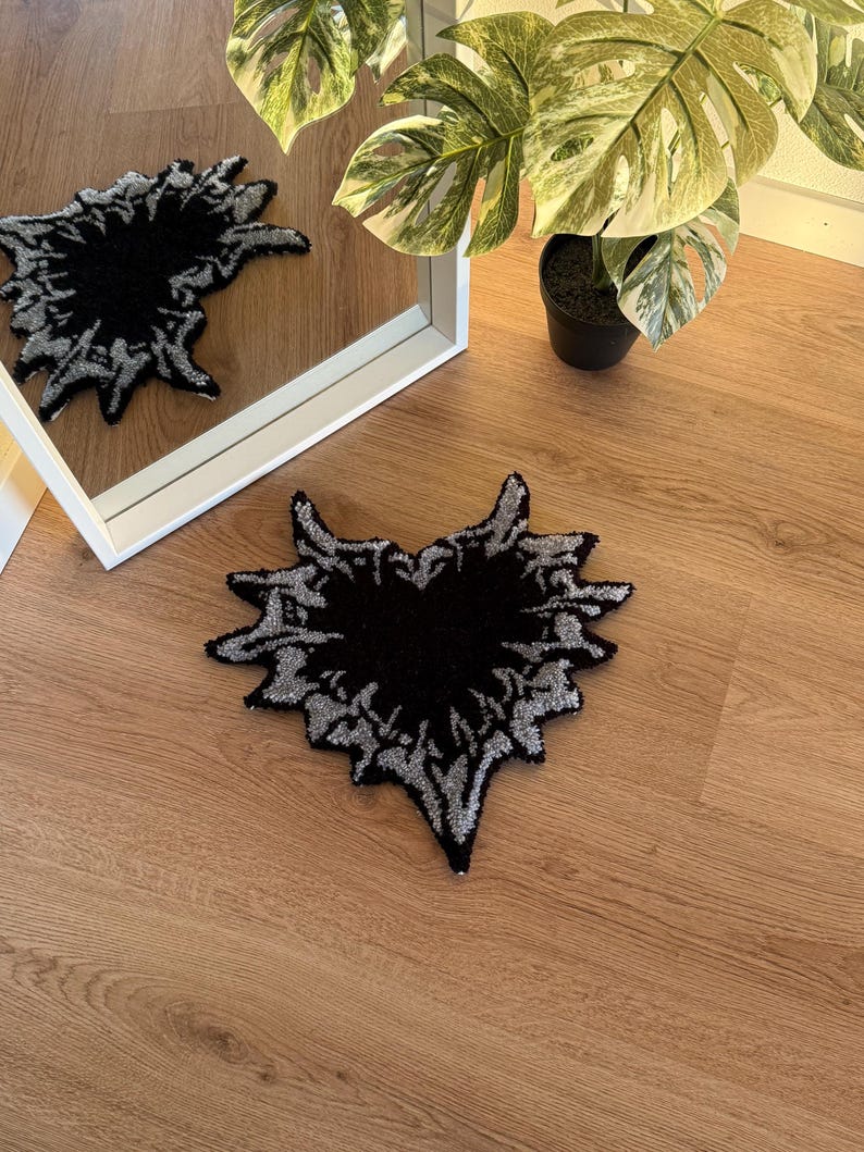 Op de afbeelding: Een zwart en zilverkleurig tapijt met een abstract ontwerp, dat lijkt op een gestileerd hart of een uitbarsting, ligt op een houten vloer. Het ontwerp van het tapijt wordt weerspiegeld in een wit omrande spiegel. Een potplant met grote, patroonbladeren staat op de achtergrond.