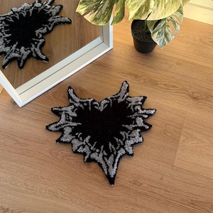Op de afbeelding: Een zwart en zilverkleurig tapijt in de vorm van een hart, met een abstract ontwerp. Het tapijt heeft een getextureerde, getufte afwerking. Het ligt op een houten vloer. Een spiegel en een plant in een pot zijn zichtbaar op de achtergrond.