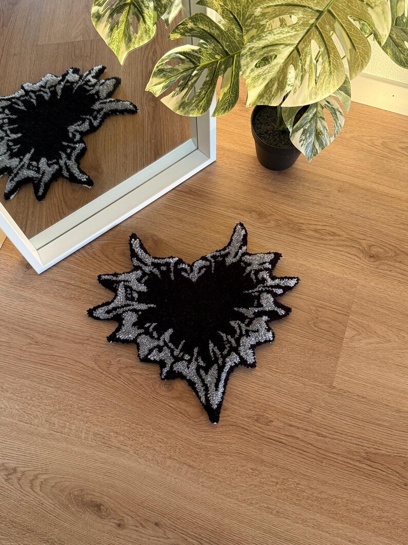 Op de afbeelding: Een zwart en zilverkleurig tapijt in de vorm van een hart, met een abstract ontwerp. Het tapijt heeft een getextureerde, getufte afwerking. Het ligt op een houten vloer. Een spiegel en een plant in een pot zijn zichtbaar op de achtergrond.