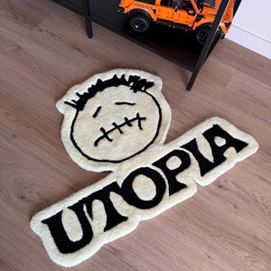 May include: A cream-colored rug with a cartoon face and the word "UTOPIA" in black. The rug has a soft, textured appearance and is placed on a wooden floor. A black shelf with a toy truck is in the background.
