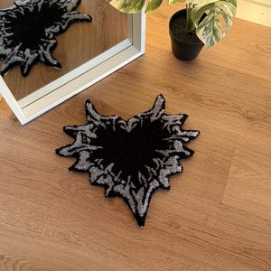 Op de afbeelding: Een zwart en zilver hartvormig tapijt met een textuurontwerp. Het tapijt ligt op een houten vloer, met een spiegel die de afbeelding van het tapijt weerspiegelt. Een plant in een pot is zichtbaar op de achtergrond.