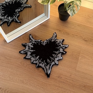 Op de afbeelding: Een zwart en zilver hartvormig tapijt met een textuurontwerp. Het tapijt ligt op een houten vloer, met een spiegel die de afbeelding van het tapijt weerspiegelt. Een plant in een pot is zichtbaar op de achtergrond.
