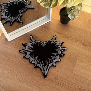 Op de afbeelding: Een zwart en zilverkleurig tapijt met een uniek, abstract ontwerp. Het tapijt heeft de vorm van een gestileerd hart of vlam, met een donker centrum en een gedetailleerde zilveren rand. Het ligt op een houten vloer.