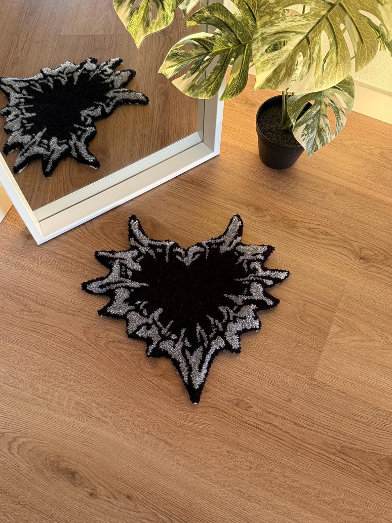 Op de afbeelding: Een zwart en zilverkleurig tapijt met een uniek, abstract ontwerp. Het tapijt heeft de vorm van een gestileerd hart of vlam, met een donker centrum en een gedetailleerde zilveren rand. Het ligt op een houten vloer.