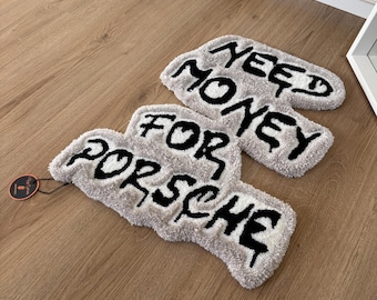 Puede incluir: Dos alfombras grises en forma de nube con texto negro "NEED MONEY FOR PORSCHE". Las alfombras están sobre un suelo de madera marrón claro, con una etiqueta adjunta a una alfombra.