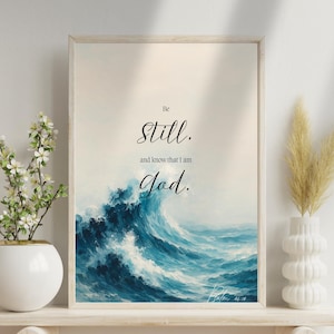 Könnte beinhalten: Ein gerahmter Kunstdruck mit einem Aquarellgemälde einer großen Meereswelle in Blautönen. Der Text "Be Still, and know that I am God" ist in eleganter Schrift dargestellt. Das Kunstwerk wird auf einem weißen Regal ausgestellt.