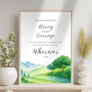 Op de afbeelding: Een ingelijste aquarelprint met de tekst: "Heb Ik u niet geboden? Wees sterk en moedig...Waar u ook gaat." Het kunstwerk toont een landschap met groene bomen, glooiende heuvels en water, in een licht houten frame.