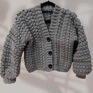 Pode incluir: Um cardigan cinza, feito à mão em crochê, com um padrão de ponto texturizado em forma de bolha. O suéter tem decote em V, mangas compridas e três botões pretos. A peça de roupa é exibida em um cabide preto sobre um fundo branco.