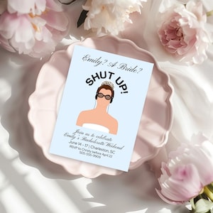 May include: A light blue invitation card with the text "Emily? A Bride? SHUT UP!" and a cartoon illustration of a woman wearing sunglasses and a tiara. The card is on a pink plate, surrounded by pink flowers.