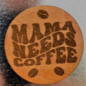 May include: A round wooden coaster with the text "Mama Needs Coffee" in brown lettering. The coaster has three coffee bean designs around the text.