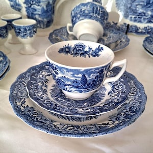 Könnte beinhalten: Ein weißes Keramik-Tassen- und Untertassen-Set mit einem blauen Transferdruck-Muster aus floralen Bordüren und einer Landschaftsszene. Die Tasse hat einen gewellten Rand mit Blumenmuster, und die Untertasse sowie der passende Teller haben einen detaillierten floralen Rand.
