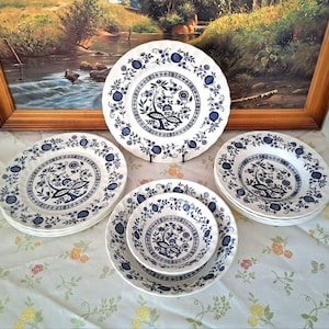 Könnte beinhalten: Eine Sammlung weißer Keramikplatten und -schalen mit blauem Blumendesign. Die Teller und Schalen sind auf einer floralen Tischdecke vor einem gerahmten Landschaftsgemälde angeordnet. Das Set umfasst Essteller, Salatteller und Schalen.