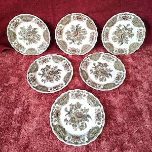 Può includere: Un set di sei piatti rotondi in ceramica con centro bianco e un motivo centrale floreale e di uccelli nei toni del rosso, giallo e blu. I bordi smerlati sono decorati con un dettagliato motivo ornamentale marrone e dorato.