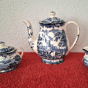 May include: A three-piece tea set featuring a teapot with a lid, a lidded sugar bowl, and a creamer. Each piece is made of cream-colored ceramic with a dark blue transferware pattern depicting a landscape with buildings, trees, and figures.