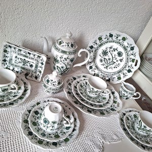 Puede incluir: Un juego de té completo de porcelana blanca con un estampado floral verde. El juego incluye tazas, platillos, platos, una tetera, un azucarero y una jarra de crema. Los platos tienen bordes festoneados. El juego de té está colocado sobre un mantel de encaje blanco.