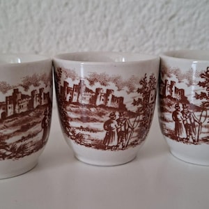 Könnte beinhalten: Drei cremeweiße Eierbecher aus Keramik mit braunem Transferdruck. Jeder Becher zeigt eine detaillierte Szene mit einer Steinburg, Bäumen und Figuren in historischer Kleidung. Das Design enthält ein kleines Schild mit einer Ente.