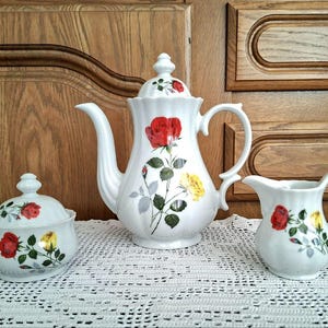 Könnte beinhalten: Ein weißes Keramik-Teeservice mit Teekanne, Sahnekännchen und Zuckerdose. Jedes Teil ist mit roten und gelben Rosenblütenmustern verziert. Das Set wird auf einer weißen Spitzendecke präsentiert.