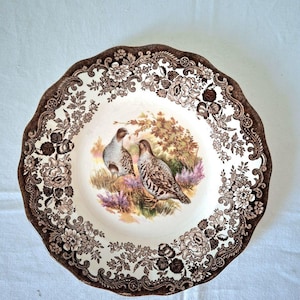 Könnte beinhalten: Eine Vintage-Keramikplatte mit einem braunen Blumenrand und einem zentralen Bild von zwei Vögeln auf einem Feld. Der Teller hat einen gewellten Rand und einen cremefarbenen Hintergrund.