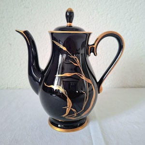 Puede incluir: Una tetera de cerámica azul oscuro con asa, boquilla y ribete dorados. La tetera presenta un diseño de espiga de trigo dorada en la parte delantera. La tapa tiene un adorno dorado. La tetera está sobre una superficie blanca.