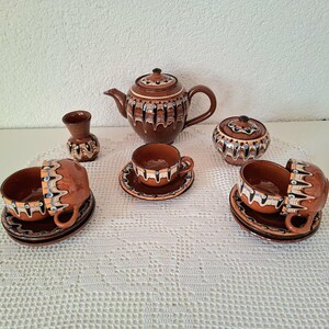 Könnte beinhalten: Ein braunes Keramik-Teeservice mit dekorativen Mustern. Das Set beinhaltet eine Teekanne, eine Zuckerdose, eine kleine Vase sowie mehrere Tassen und Untertassen. Die Tassen und Untertassen haben ein einzigartiges Griffdesign. Das Set wird auf einer weißen Spitzendecke präsentiert.
