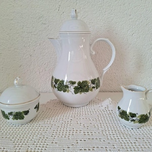 Könnte beinhalten: Weißes Porzellan-Teeservice mit grünem Blattmuster. Das Set beinhaltet eine Kaffeekanne mit Deckel, eine Zuckerdose mit Deckel und einen Sahnekännchen. Goldfarbene Verzierungen akzentuieren die Ränder jedes Stücks.