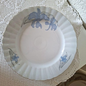 Könnte beinhalten: Weißer Keramikteller mit gewelltem Rand und Goldrand. Der Teller ist mit blauen Blumendesigns verziert. Der Teller liegt auf einer weißen Spitzendecke.