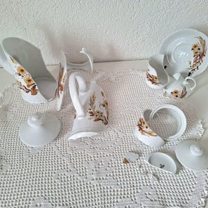 Könnte beinhalten: Weißes Porzellan-Teeservice mit floralem Design in Braun- und Gelbtönen. Das Set umfasst eine Teekanne, einen Sahnekännchen, eine Zuckerdose, Tassen, Untertassen und ein Sieb. Die Teile sind auf einer weißen Häkeldecke angeordnet.