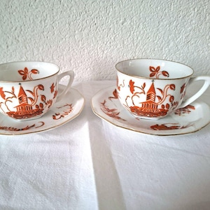 Könnte beinhalten: Zwei weiße Tassen und Untertassen mit orangefarbenen und goldenen Akzenten. Jede Tasse und Untertasse zeigt eine detaillierte orangefarbene Illustration eines Gebäudes und floraler Elemente. Die Ränder sind goldfarben.