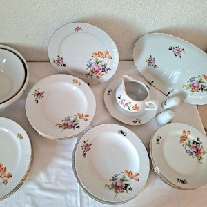 Peut inclure: Service de table en porcelaine blanche avec un motif floral. L'ensemble comprend des assiettes, des bols, un plat de service, une crémière et des salières et poivrières. Le motif floral présente des fleurs roses, violettes, oranges et bleues avec une bordure dorée.