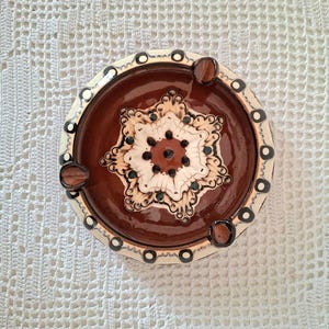 Könnte beinhalten: Ein runder Aschenbecher aus Keramik mit braunem Innenraum und cremefarbenem Rand. Die Mitte zeigt ein weißes und braunes Blumenmuster. Der Rand hat ein sich wiederholendes Muster aus schwarzen und cremefarbenen Kreisen. Drei kleine Ablagen sind am Rand angebracht.