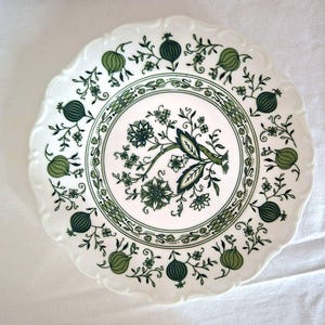 Puede incluir: Plato de cerámica blanca con borde festoneado, con un diseño floral y frutal verde. El centro tiene un arreglo floral detallado, rodeado por un borde decorativo y motivos de frutas repetidos. Un plato clásico de estilo vintage.