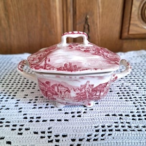 Può includere: Una zuppiera in ceramica con coperchio e un motivo a paesaggio rosso e bianco. La zuppiera ha un coperchio con manico, due manici laterali e tre piedini. È appoggiata su una tovaglia di pizzo bianco.