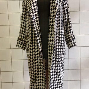 May include: A long, black and white houndstooth pattern coat. The coat is displayed on a mannequin. The coat has a classic design with a notched collar, button closure, and long sleeves. The coat's length extends to the ankles.