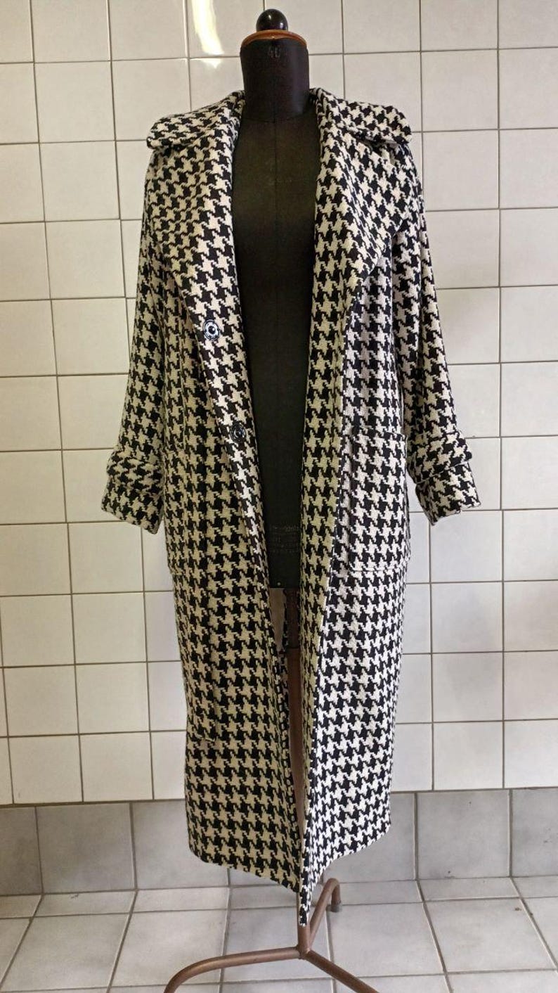 May include: A long, black and white houndstooth pattern coat. The coat is displayed on a mannequin. The coat has a classic design with a notched collar, button closure, and long sleeves. The coat's length extends to the ankles.