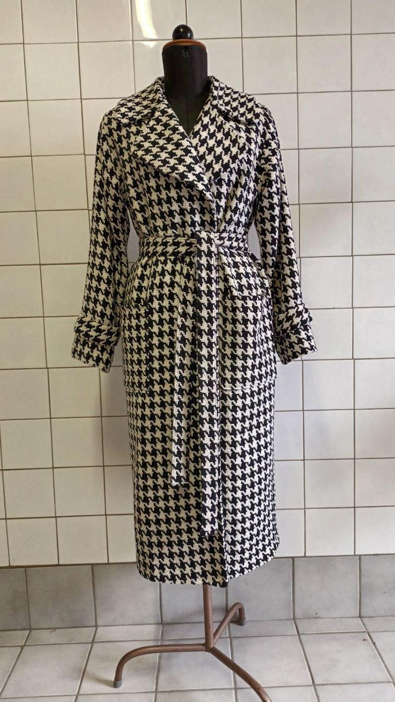 May include: A long, houndstooth-patterned coat in black and cream, displayed on a mannequin. The coat features a wide collar, long sleeves, a belt, and pockets. The coat is knee-length and has a classic, timeless design.