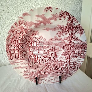 Könnte beinhalten: Dekorative Platte mit rot-weißem Transferdruck. Die Platte zeigt eine ländliche Szene mit einer Pferdekutsche, Gebäuden und Bäumen. Die Platte wird von einem schwarzen Ständer gehalten.