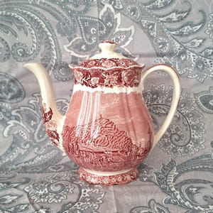 Könnte beinhalten: Eine Vintage-Teekanne aus rot-weißem Keramik mit Deckel. Die Teekanne hat ein Landschaftsdesign und florale Akzente. Der Griff und der Ausguss sind weiß. Die Teekanne steht auf einer grau-weißen Paisley-Oberfläche.