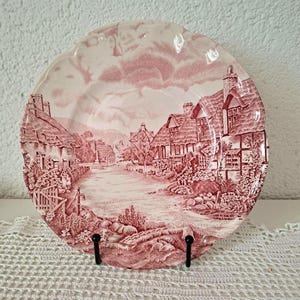 Puede incluir: Plato decorativo con borde festoneado, con una escena pastoral roja y blanca. El diseño representa un pueblo con casas, un río y árboles. El plato se exhibe sobre un soporte negro sobre un fondo blanco.