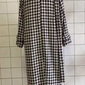 May include: A long, houndstooth-patterned coat in black and white. The coat has long sleeves with folded cuffs and a classic collar. The coat is displayed on a mannequin against a white tiled wall.