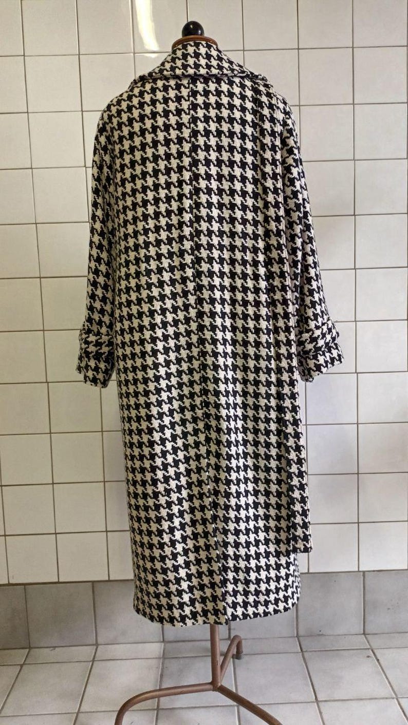 May include: A long, houndstooth-patterned coat in black and white. The coat has long sleeves with folded cuffs and a classic collar. The coat is displayed on a mannequin against a white tiled wall.