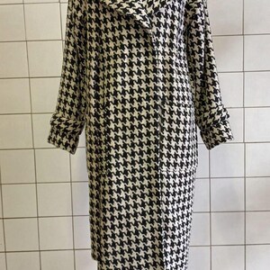 May include: A black and white houndstooth pattern coat displayed on a mannequin. The long coat has a notched collar, long sleeves, and pockets. The coat is knee-length and has a classic, timeless design.
